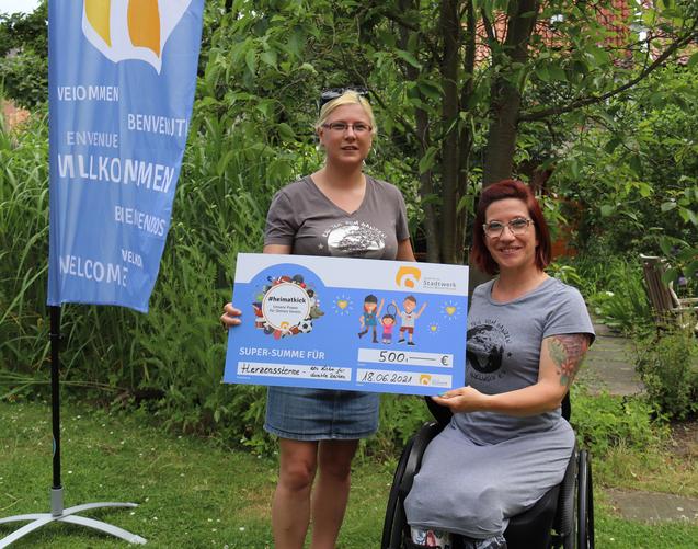 Zwei Frauen halten einen Scheck &uuml;ber 500 Euro f&uuml;r ein Projekt, umgeben von gr&uuml;ner Natur und einem Willkommensbanner.