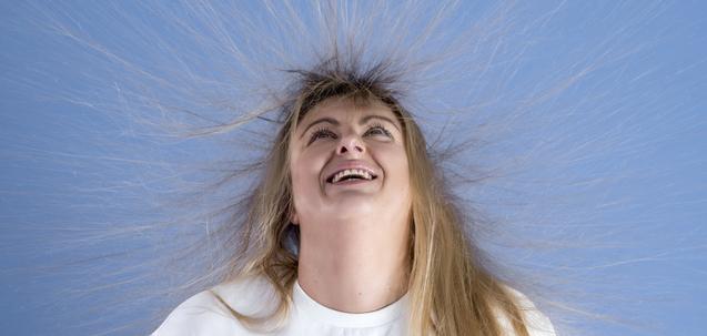 Frau mit strähnigem Haar lächelt auf blauem Hintergrund, der Haar steht aufgrund statischer Aufladung ab.