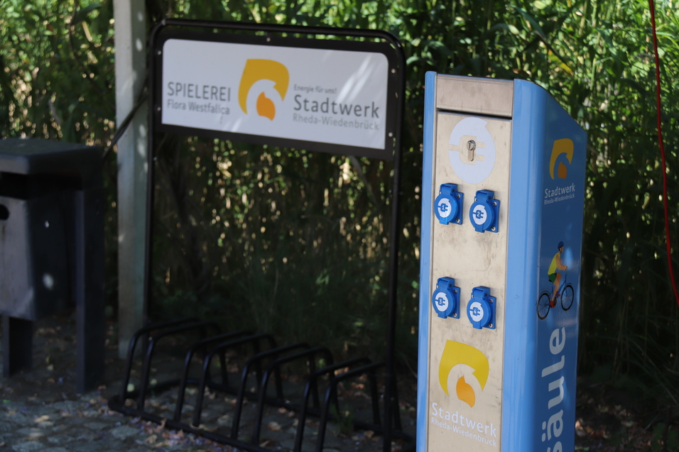 Ladesäule für Elektrofahrzeuge mit Steckdosen und einem Fahrradständer im Hintergrund in einer parkähnlichen Umgebung.