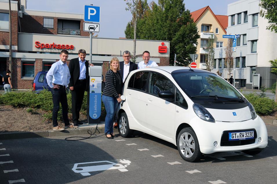 Fünf Personen stehen neben einem weißen Elektroauto, das an einer Ladestation aufgeladen wird, vor einer Sparkasse.