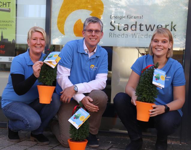 Drei Personen in blauen T-Shirts halten kleine Tannenb&auml;ume in T&ouml;pfen vor einem Stadtwerk-Logo.