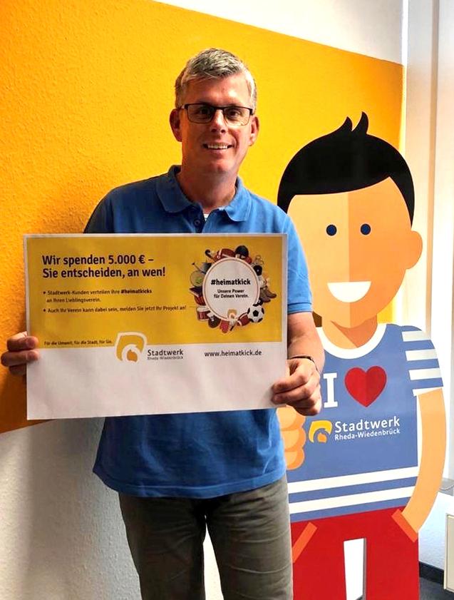 Mann h&auml;lt ein Plakat mit der Aufschrift &bdquo;Wir spenden 5.000 &euro; &ndash; Sie entscheiden, an wen!&ldquo; vor einer Wand mit einem Comic-Figur.